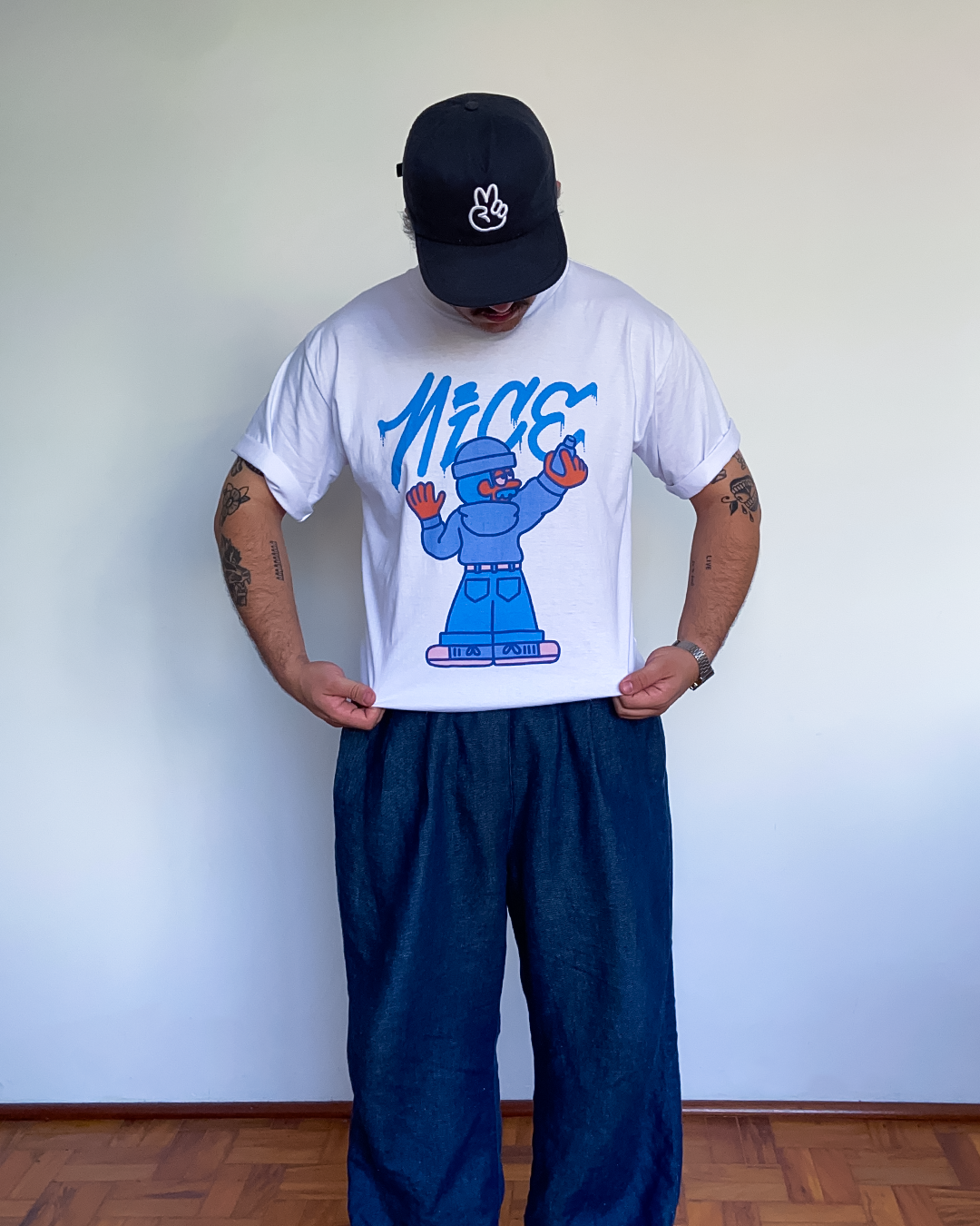 White El Cabriton T-shirt worn with blue jeans, featuring a colorful character painting the word Nice.